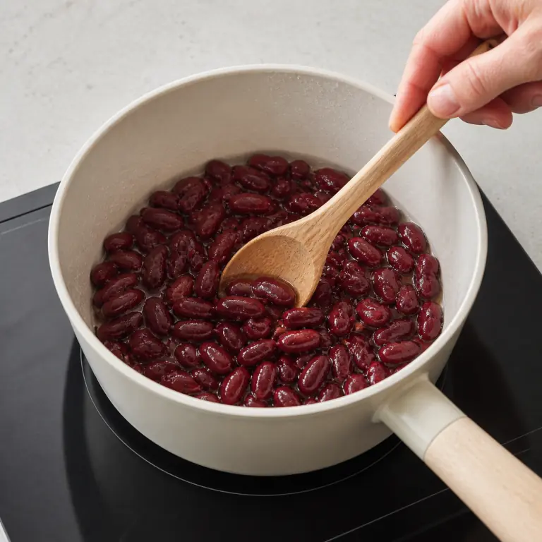 Gros plan pédagogique de réchauffe doucement 80 g de haricots rouges sans les écraser. Montrer le récipient exact, une main adulte avec l'ustensile adapté et la couleur ou texture qui prouve la cuisson. Aucun autre aliment ne doit masquer le résultat.