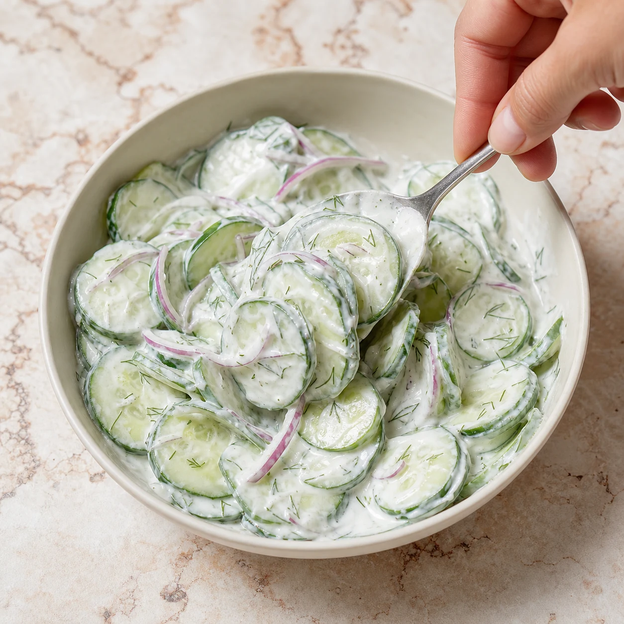 Cuillère soulevant doucement des rondelles de concombre nappées de sauce blanche à l'aneth et à l'oignon rouge dans un bol.