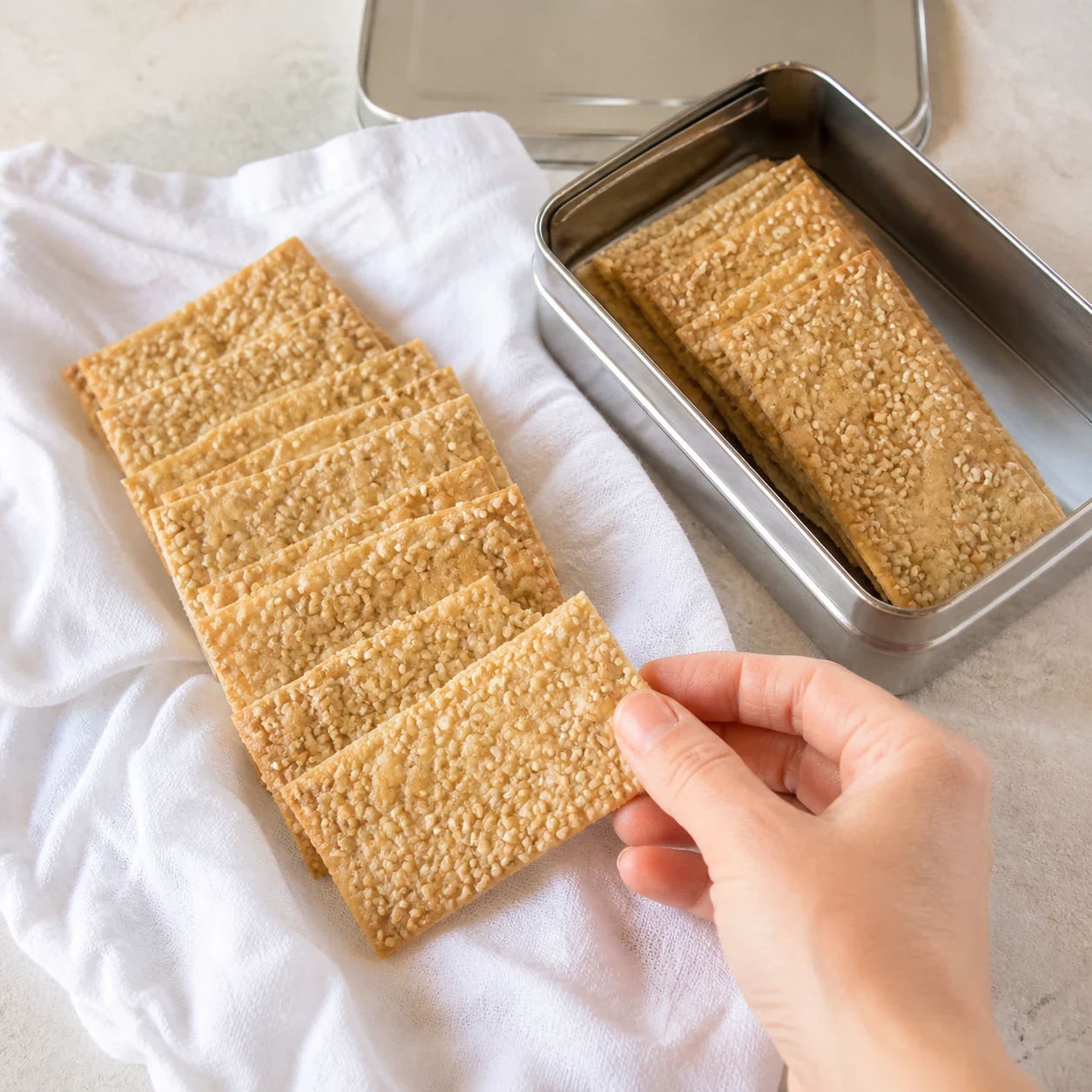 Crackers rectangulaires beige doré séparés et empilés en éventail sur un torchon en coton blanc, surface constellée de graines de sésame, une boîte en métal ouverte à côté, aucun romarin, aucune tartinade, aucun autre ingrédient, aucun texte, aucune marque.