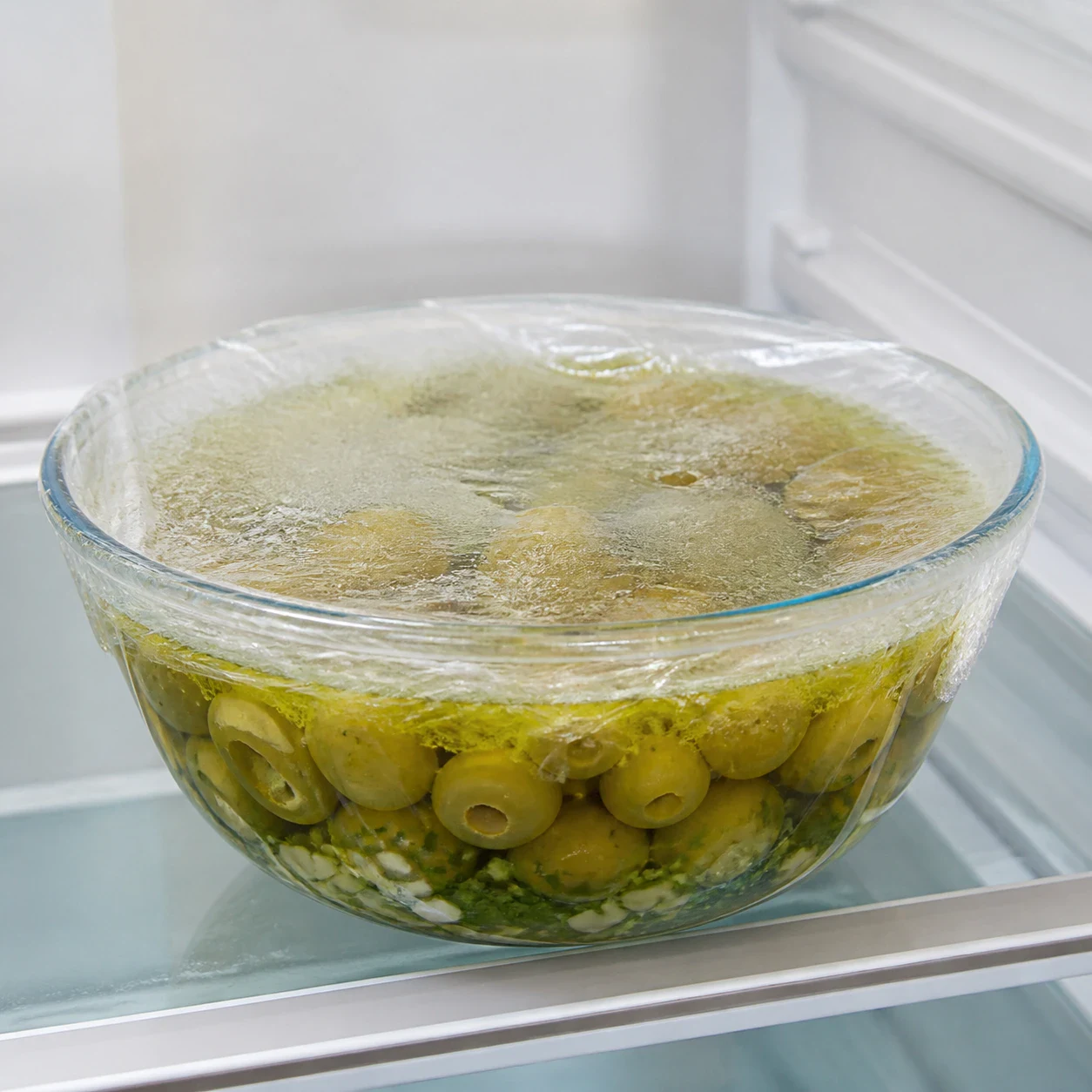 Bol en verre rempli d'olives vertes marinées à l'huile d'olive avec ail et basilic, recouvert d'un film transparent tendu, posé sur une clayette de réfrigérateur, aucune main, aucun texte, aucun filigrane, aucune marque.