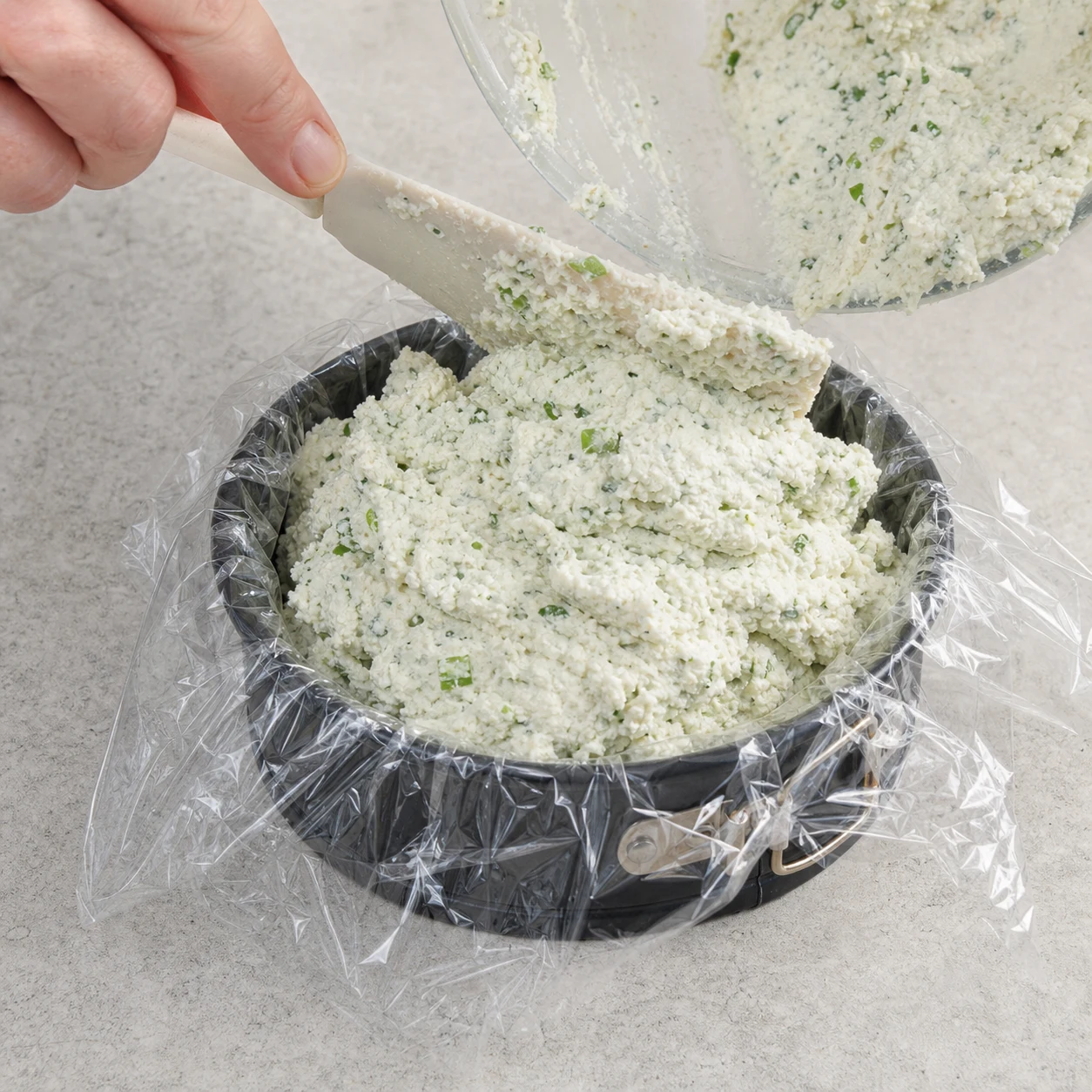 Une spatule transfère la préparation blanche aux éclats verts dans un petit moule à charnière chemisé de film alimentaire, sur plan de travail pierre pâle.