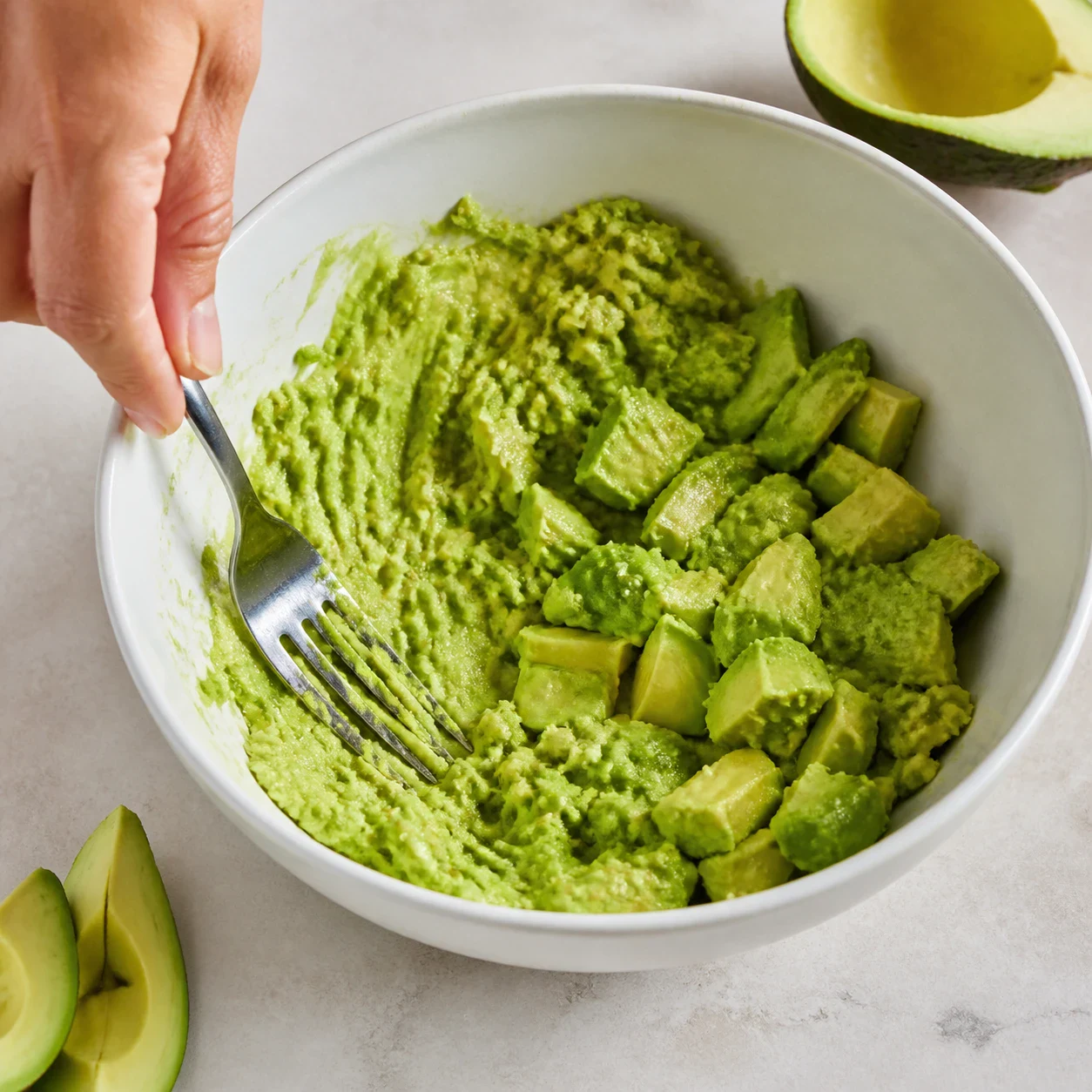 Guacamole vert en cours d'écrasement à la fourchette dans un bol blanc, purée d'un côté et morceaux d'avocat encore entiers de l'autre, fourchette appuyée contre la paroi, aucun texte, aucune marque.