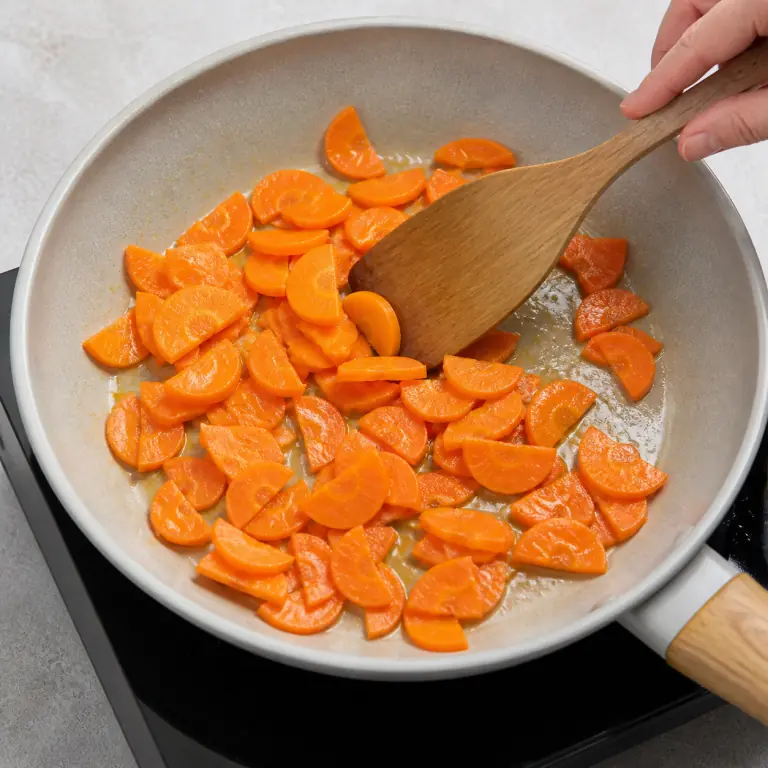 Gros plan pédagogique de fais revenir les demi-rondelles de 150 g de carotte jusqu'à ce qu'elles soient tendres mais encore nettes. Montrer le récipient exact, une main adulte avec l'ustensile adapté et la couleur ou texture qui prouve la cuisson. Aucun autre aliment ne doit masquer le résultat.