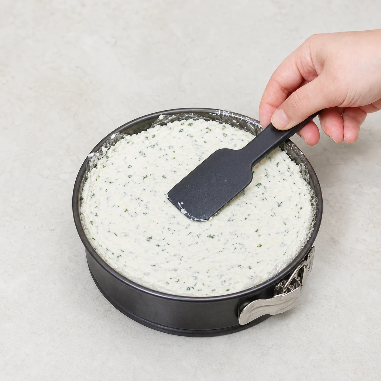 Une spatule lisse la surface blanche épaisse, parsemée d’herbes vertes, dans un moule rond chemisé ; seule une main adulte anonyme est visible.
