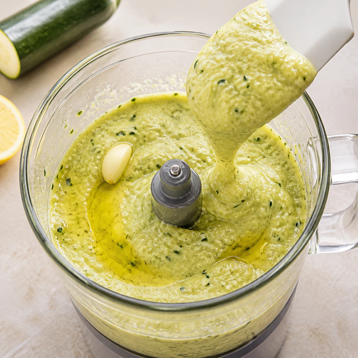 Purée de courgette vert pâle en cours de mixage dans un bol de mixeur en verre, petits éclats de peau vert foncé visibles, une demi-gousse d'ail et un filet d'huile d'olive au-dessus, aucune main, aucun texte, aucune marque.