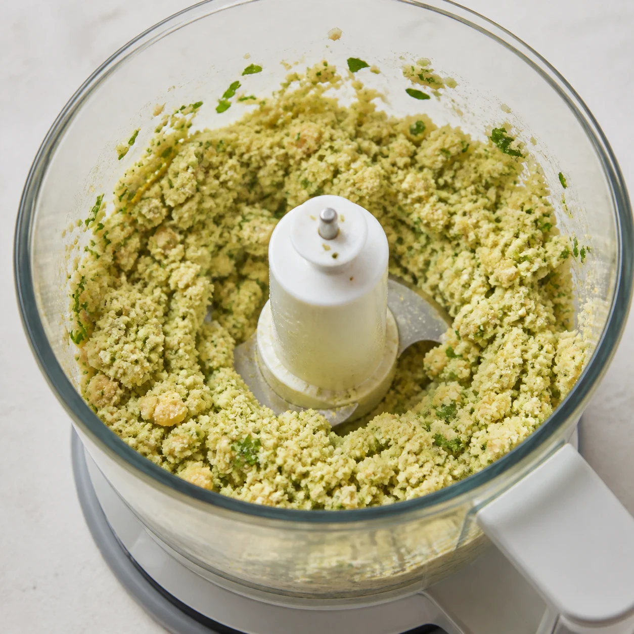 Bol de mixeur en verre contenant des pois chiches cuits, de la coriandre fraîche hachée, des morceaux d'oignon cru et une pincée de cumin moulu, texture granuleuse en cours de mixage. Aucune main, aucun texte, aucune marque.