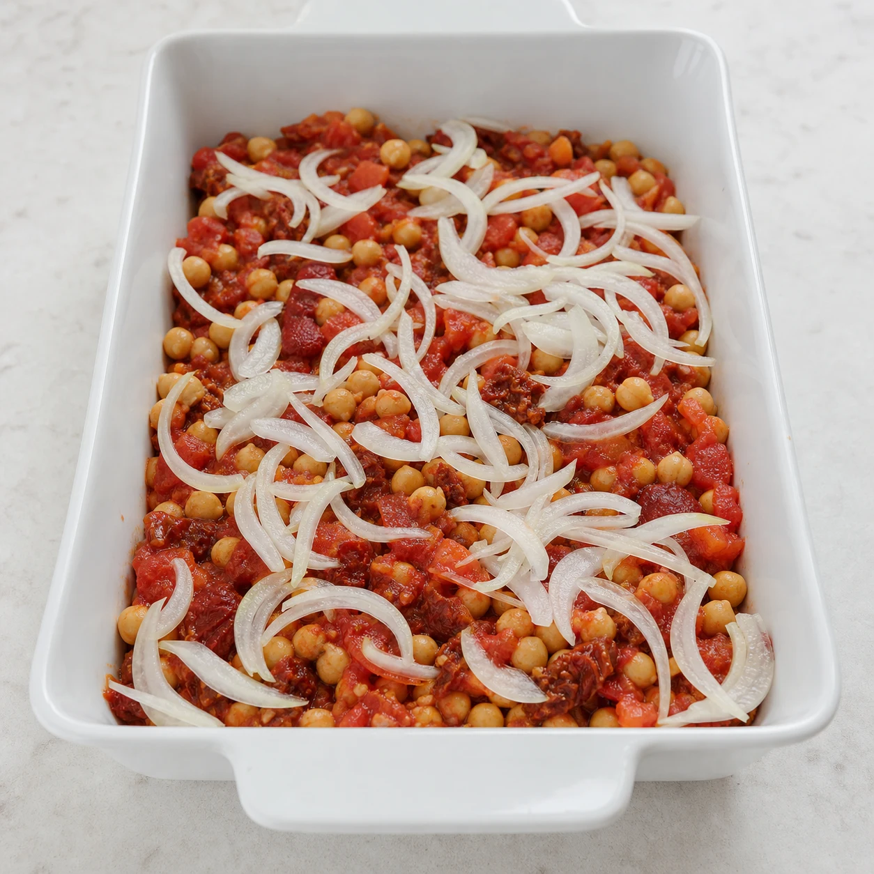 Lamelles d’oignons pâles en cours de répartition au-dessus des pois chiches et des tomates.