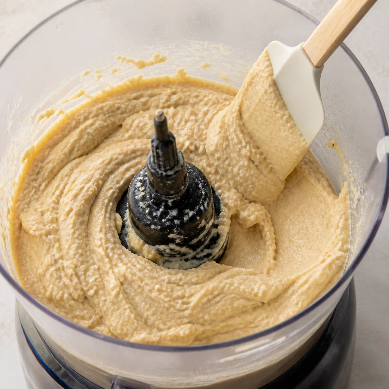 Houmous beige clair en cours de mixage dans un bol de blender, texture crémeuse avec quelques grains visibles, spatule appuyée contre la paroi, aucune main, aucun texte.