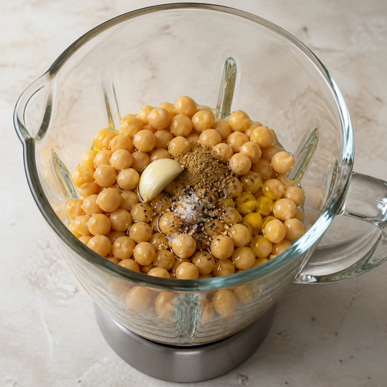 Bol de blender en verre vu de trois quarts contenant pois chiches cuits, un filet d'huile de sésame ambrée, un filet d'huile d'olive, une demi-gousse d'ail et une pincée de cumin en poudre, aucune main, aucun texte, aucune marque.