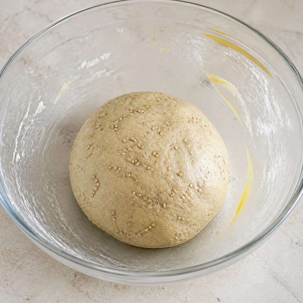 Saladier en verre contenant une boule de pâte à crackers beige clair mouchetée de graines de sésame, un peu de farine de blé et un filet d'huile d'olive encore visibles sur le bord, aucun autre ingrédient, aucun texte, aucune marque.