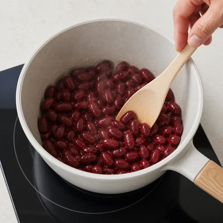 Gros plan pédagogique de réchauffe doucement 80 g de haricots rouges sans les écraser. Montrer le récipient exact, une main adulte avec l'ustensile adapté et la couleur ou texture qui prouve la cuisson. Aucun autre aliment ne doit masquer le résultat.
