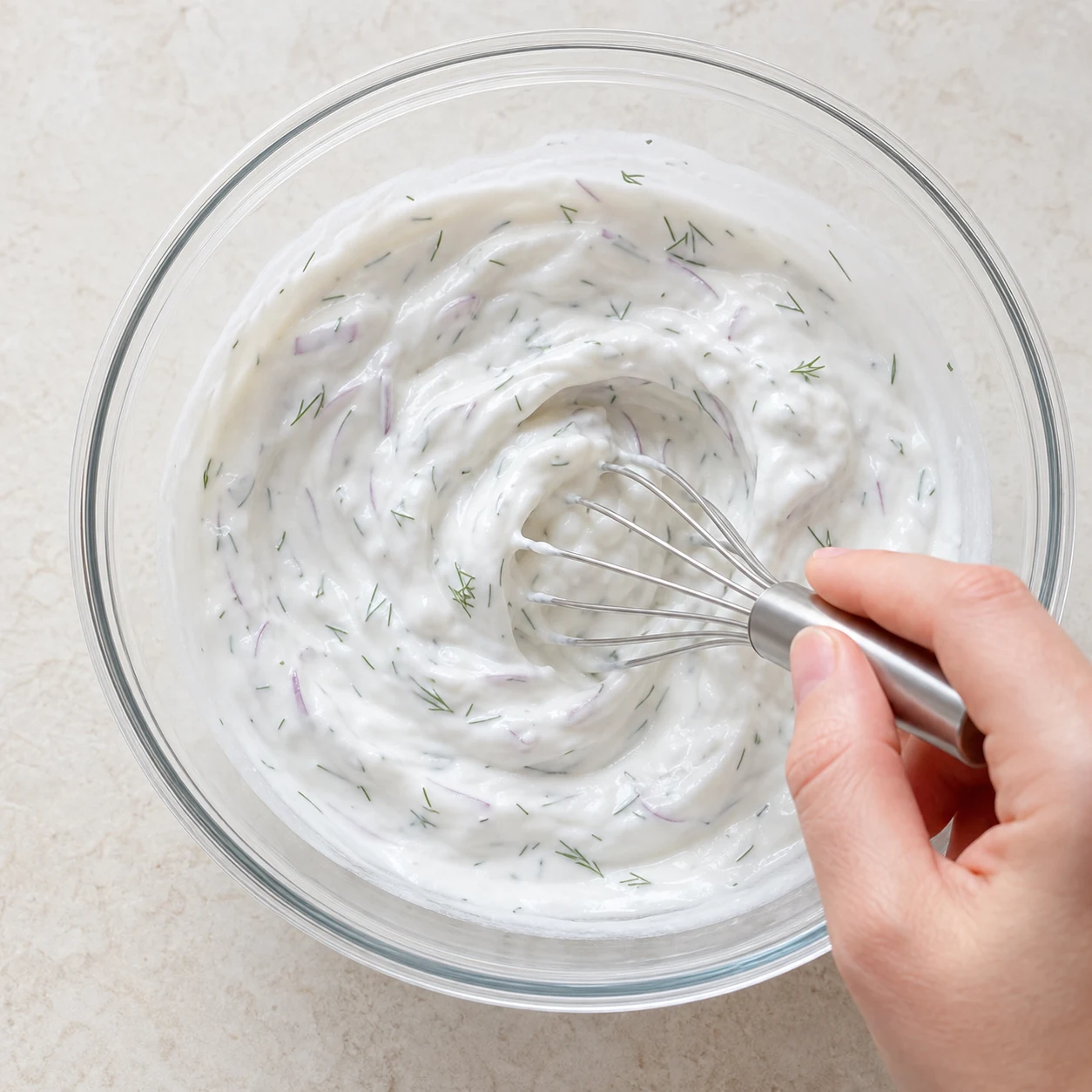 Fouet mélangeant la sauce blanche avec fines lamelles d'oignon rouge et brins d'aneth visibles, sans ingrédients futurs ajoutés.