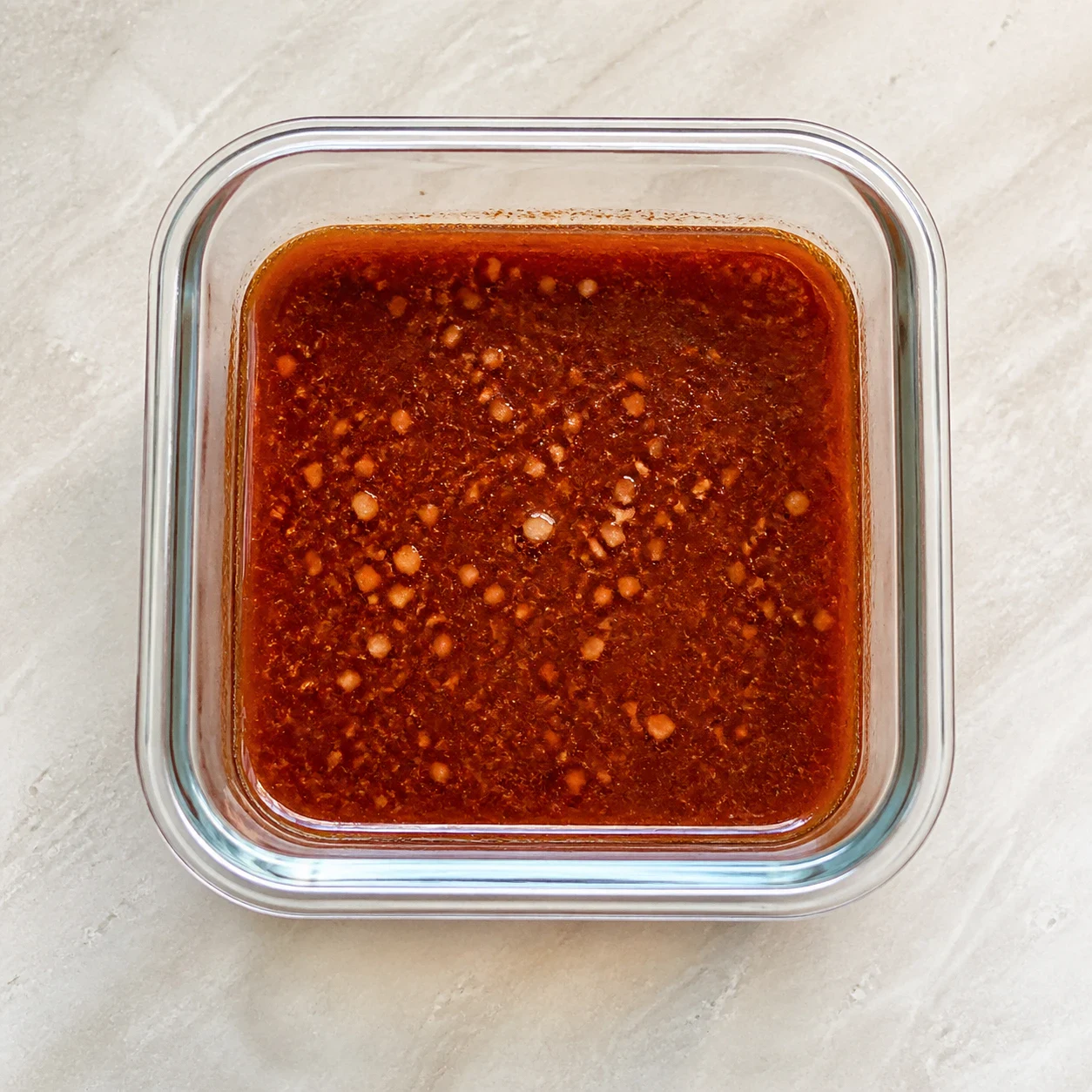 Boîte hermétique en verre ouverte vue de dessus contenant une marinade rouge brique de sauce soja, huile d'olive, paprika fumé, sirop d'érable et jus de citron, avec de l'ail pressé visible en surface, aucune main, aucun texte, aucune marque.