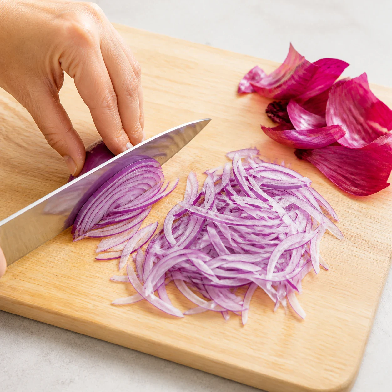 Demi-oignon rouge émincé en très fines lamelles violettes sur une planche à découper en bois clair, la pelure sèche repoussée sur le côté, aucune main, aucun autre ingrédient, aucun texte.
