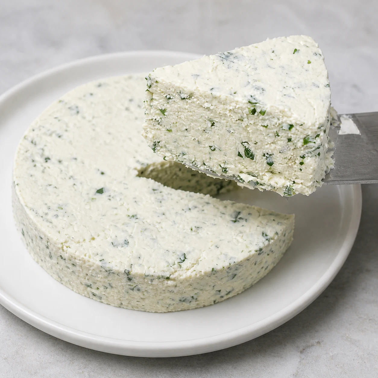 Gros plan d’une tranche nette de fromage frais végétal blanc aux éclats de persil et d’oignon nouveau, soulevée par une spatule au-dessus du fromage rond ; sans crackers, sans personne ni texte.