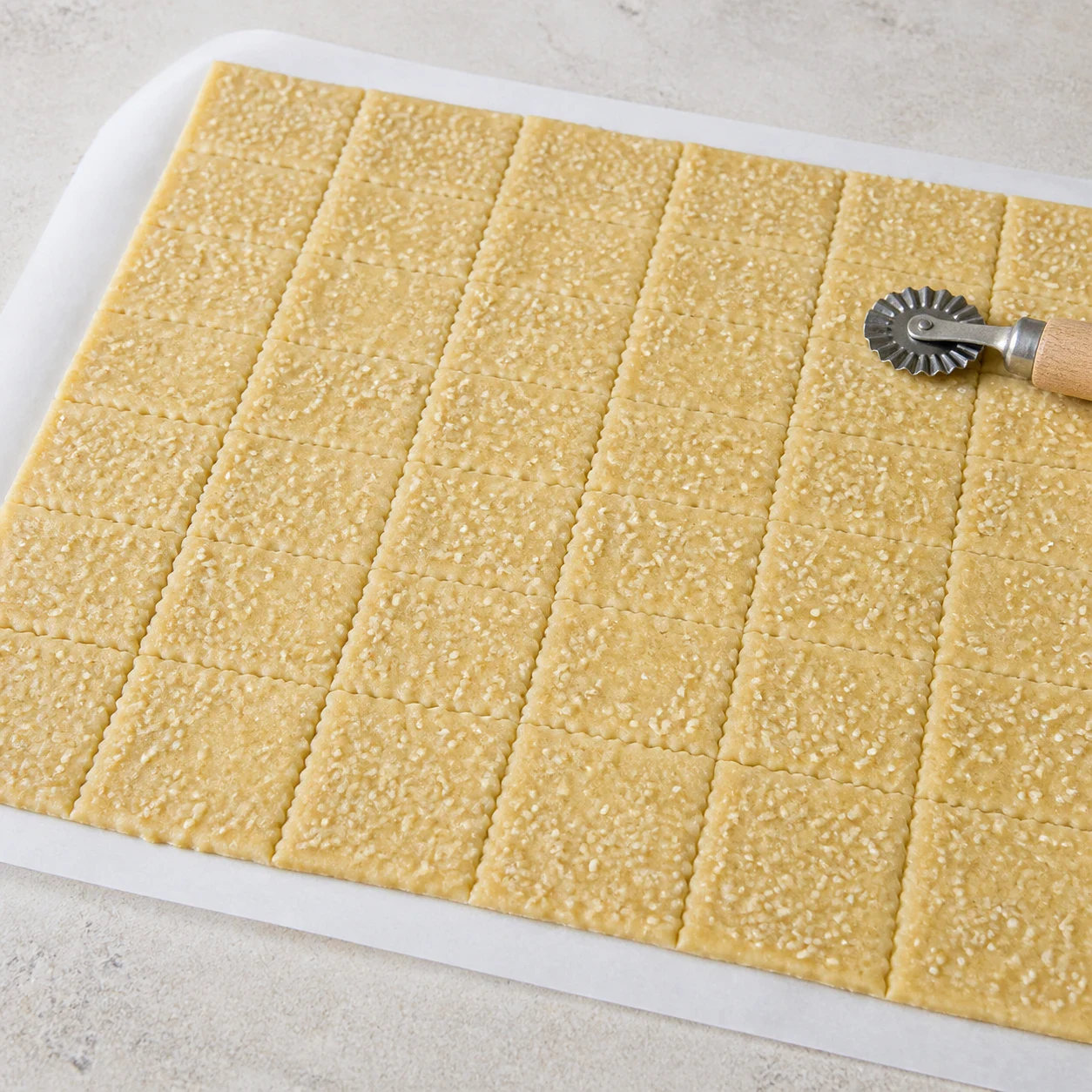 Abaisse de pâte à crackers parsemée de graines de sésame sur du papier sulfurisé, quadrillage de rectangles marqué en creux à la roulette à raviolis, roulette posée au bord de l'abaisse, aucun autre ingrédient, aucun texte, aucune marque.