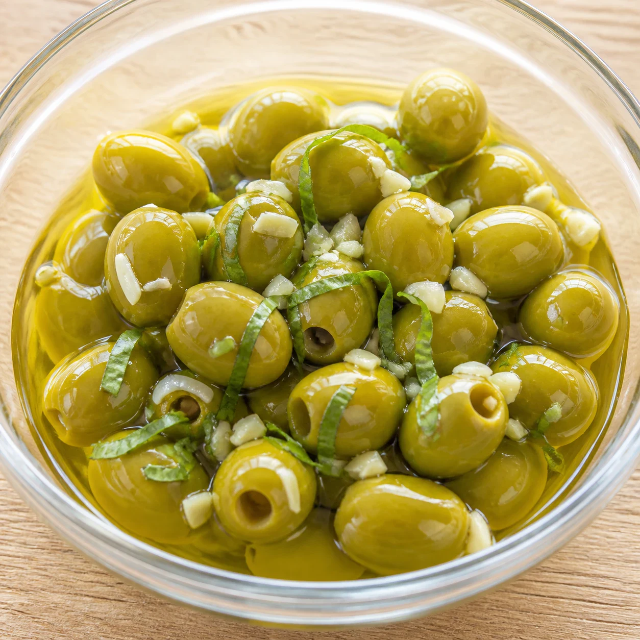 Olives vertes marinées brillantes d'huile d'olive dans un bol en verre transparent, parsemées de petits éclats d'ail blanc et de fines lanières de basilic frais, posé sur une table en bois clair, gros plan. Aucune olive noire, aucune autre herbe, aucune main, aucun texte, aucun filigrane, aucune marque.
