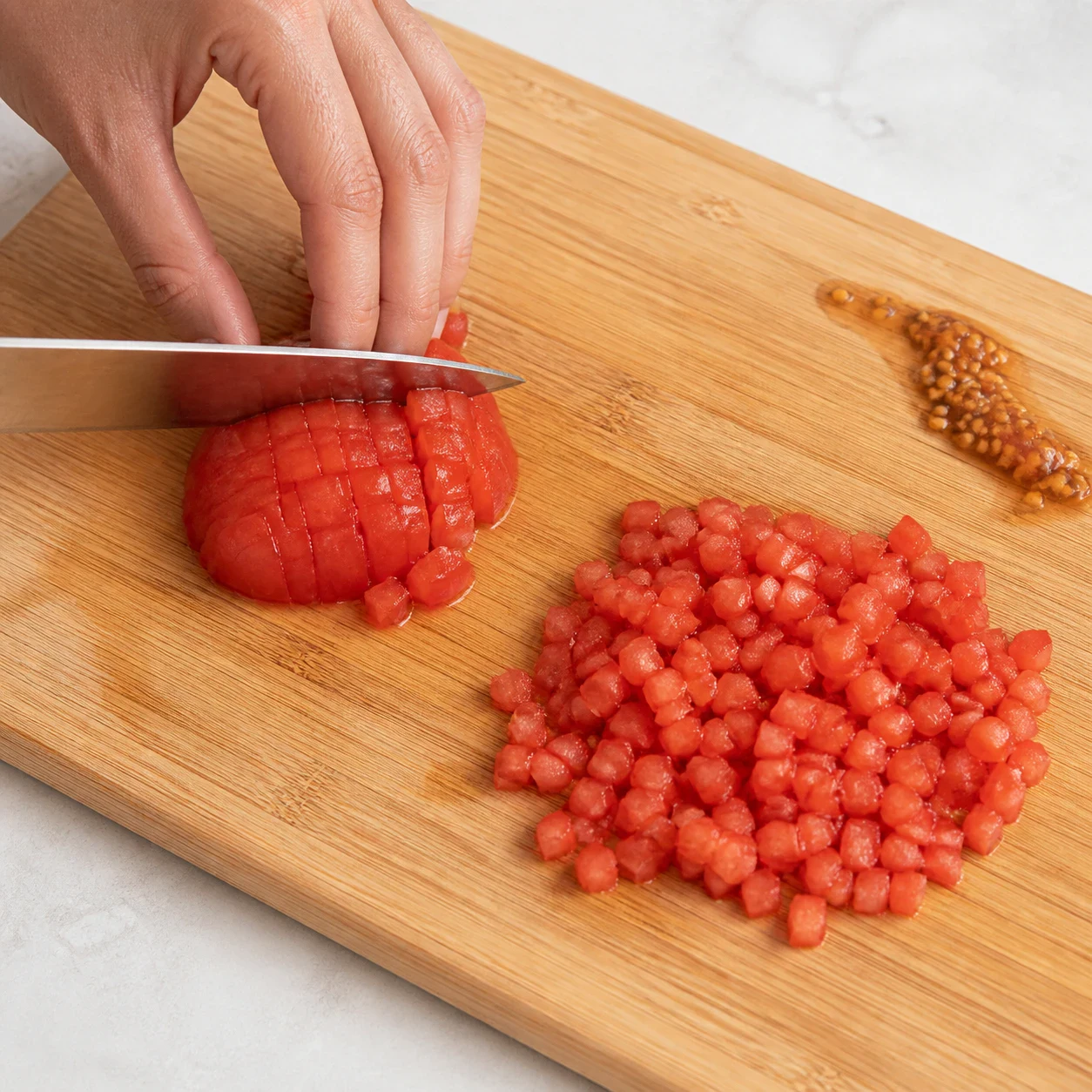 Tomate rouge en cours de découpe en petits dés sur une planche à découper en bois clair, lame du couteau visible, une main d'adulte propre tenant la tomate en griffe avec les doigts repliés, quelques dés déjà taillés à côté du morceau entier, aucun autre ingrédient, aucun texte.
