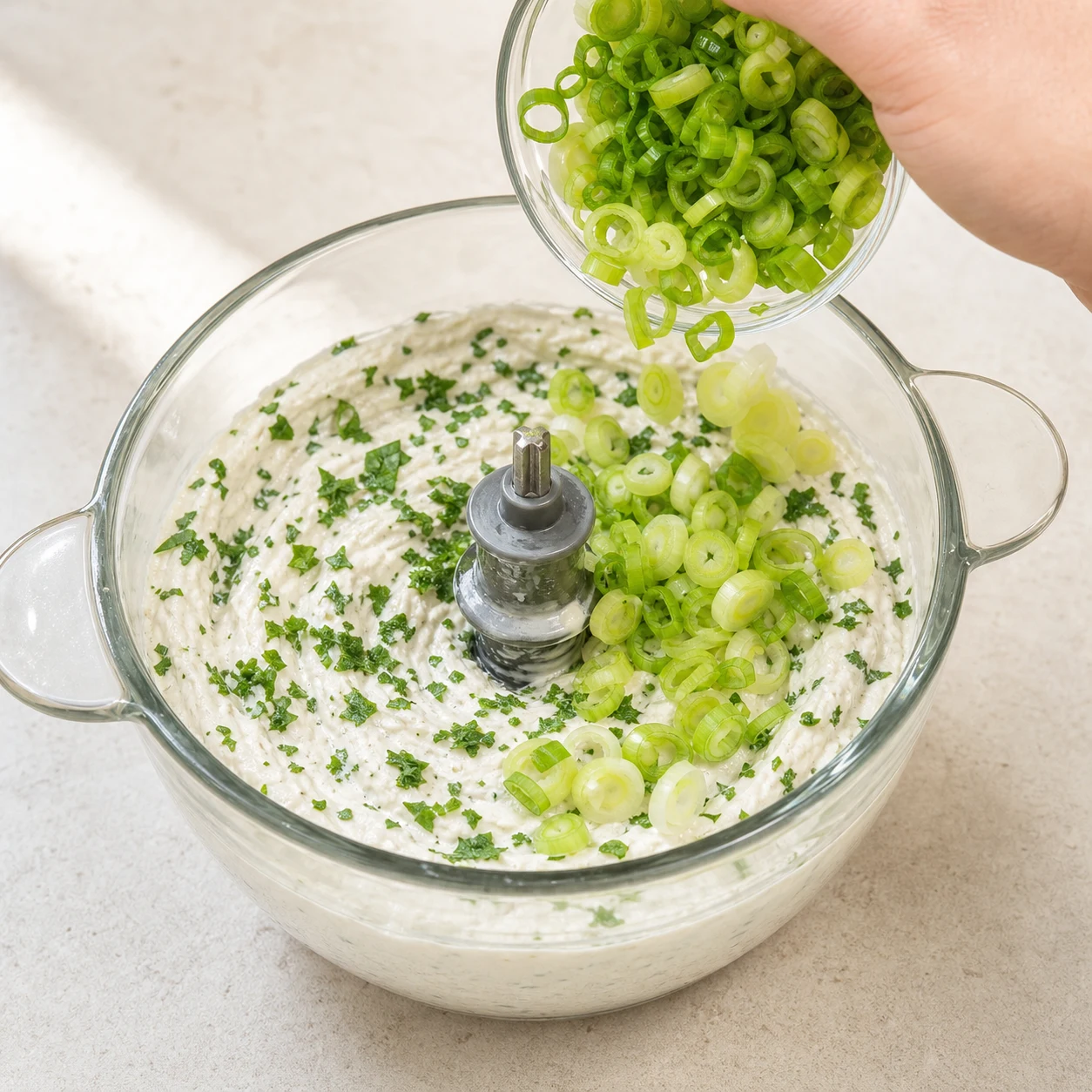 Vue rapprochée de la base blanche dans le mixeur avec persil haché et rondelles vert pâle d’oignon nouveau visibles ; aucun geste non sourcé n’est suggéré.