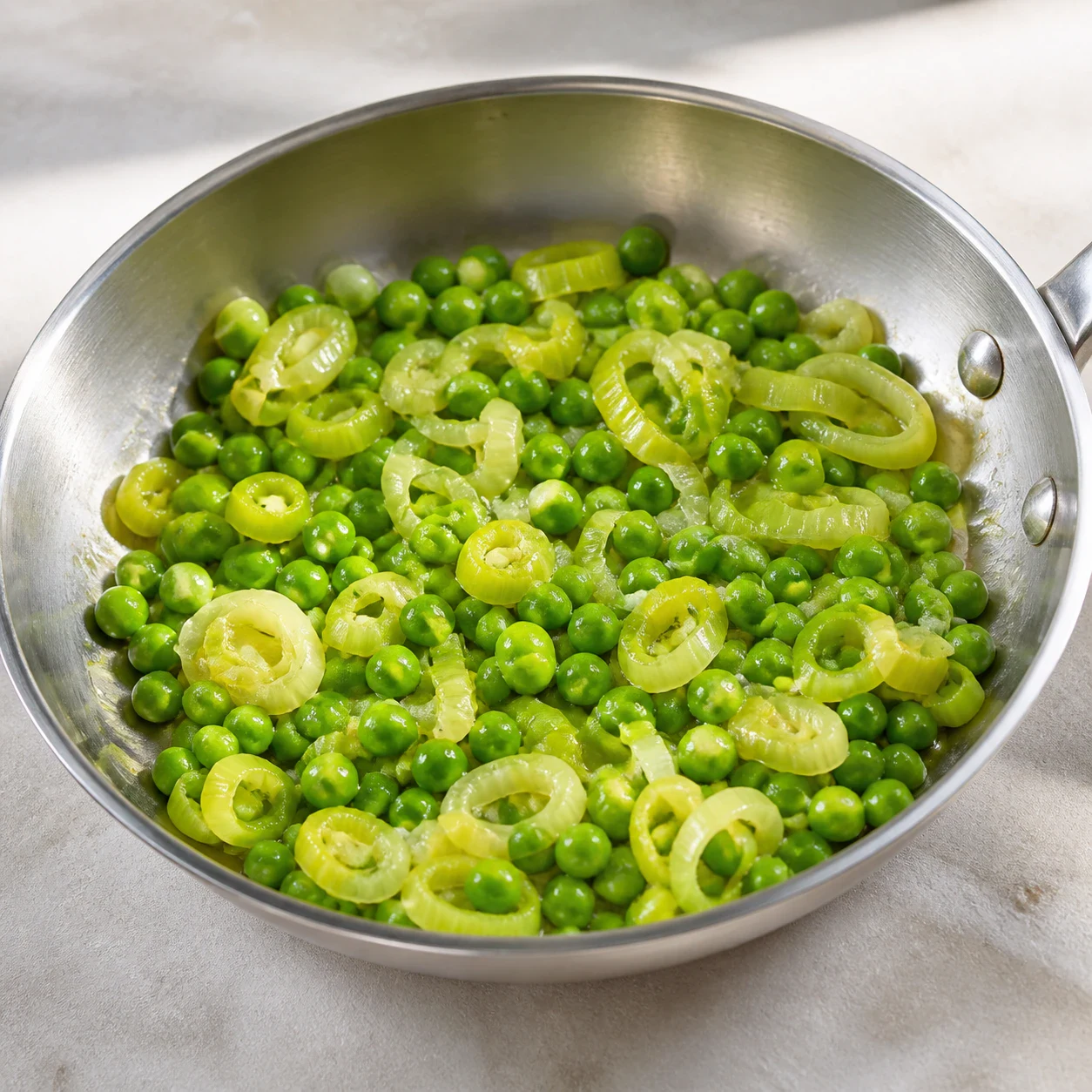 Poêle en inox contenant des morceaux de poireau devenus translucides et des petits pois vert vif, brillants d'huile d'olive. Aucune main, aucun texte, aucune marque.