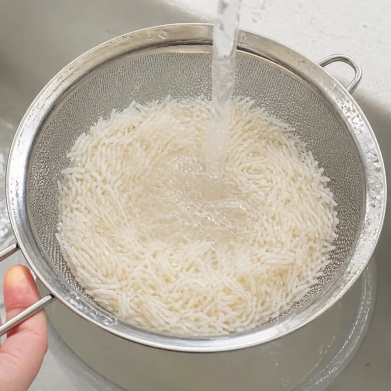 Gros plan pédagogique du geste en cours : rince 80 g de riz basmati dans une passoire fine jusqu'à ce que l'eau soit presque claire. Montrer uniquement cet ingrédient et l'ustensile nécessaire.