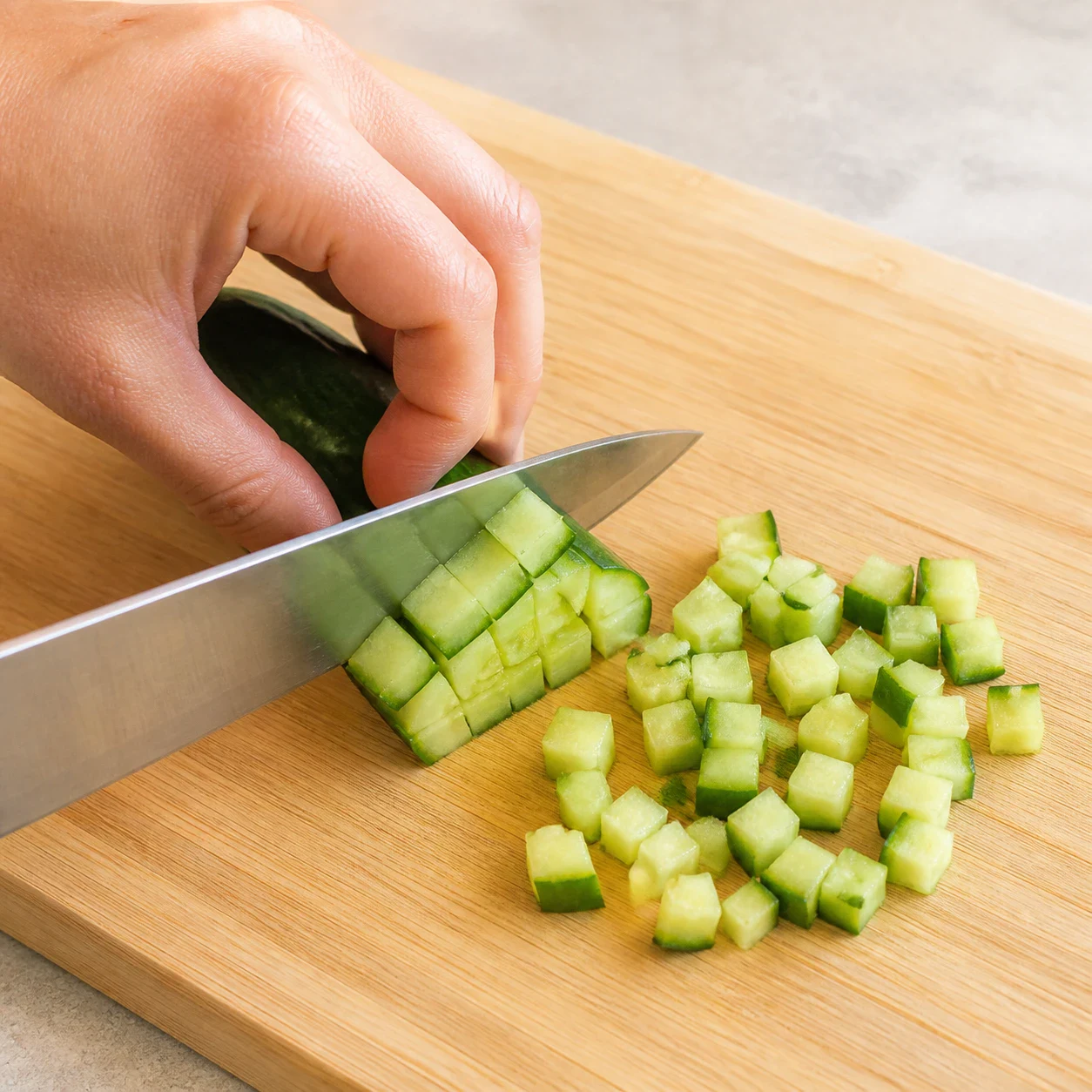 Demi-concombre vert en cours de découpe en petits dés sur une planche à découper en bois clair, lame du couteau visible, une main d'adulte propre tenant le concombre en griffe avec les doigts repliés, quelques dés déjà taillés à côté, aucun autre ingrédient, aucun texte.