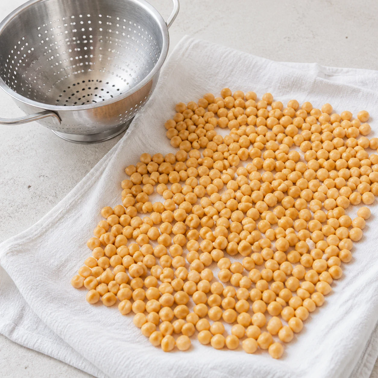 Pois chiches cuits égouttés étalés sur un linge de cuisine blanc, grains mats et secs, passoire métallique vide posée à côté, lumière naturelle. Aucun autre ingrédient, aucune personne, aucun texte.